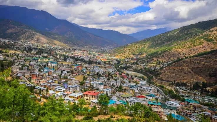 Panoramic high-angle view of Thimphu Valley in Bhutan, showcasing a sprawling mountain city nestled between steep green ridges under a partly cloudy sky.